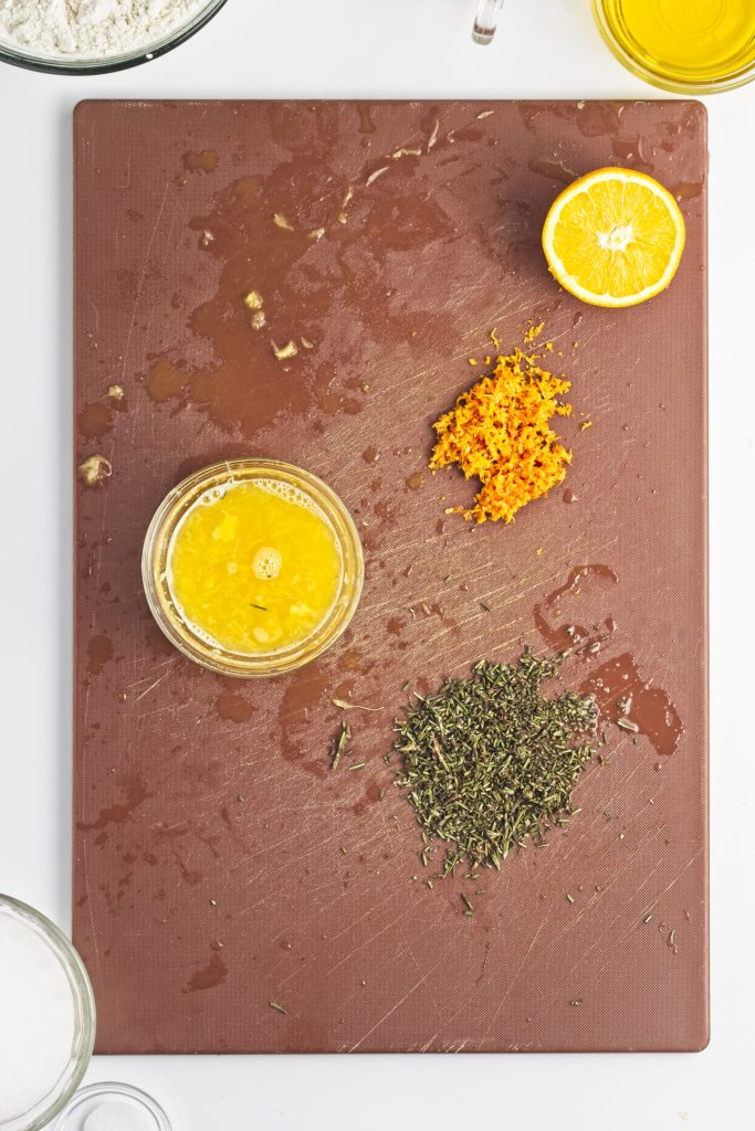 A cutting board with a bowl of orange juice, orange zest, and chopped fresh rosemary. Surrounding the bowl are various other glass bowls filled with ingredients ready to mix.