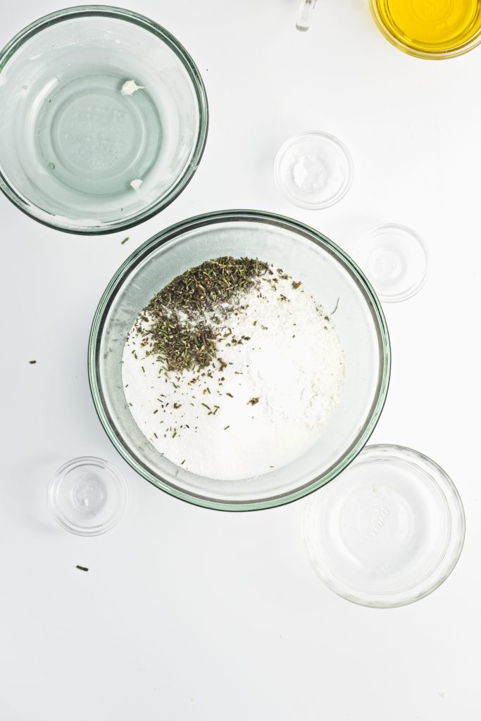 The dry ingredients for the olive oil orange cake are in a glass bowl ready to mix. Several empty smaller glass bowls surround it. There is a bowl with olive oil in the top right corner.
