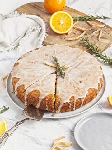 Vegan olive oil orange and rosemary cake on a white plate with orange halves and fresh sprigs of rosemary on top with a slice cut and being lifted with a cake server.