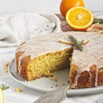 Vegan olive oil orange and rosemary cake on a white plate with orange halves and fresh sprigs of rosemary on top with a slice on a smaller plate and a fork and cake server.