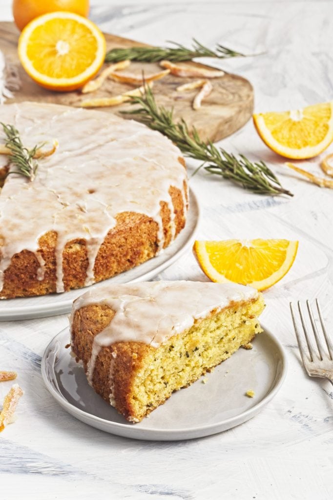 Vegan olive oil orange and rosemary cake on a white plate with orange halves and fresh sprigs of rosemary on top with a slice on a smaller plate and a fork.