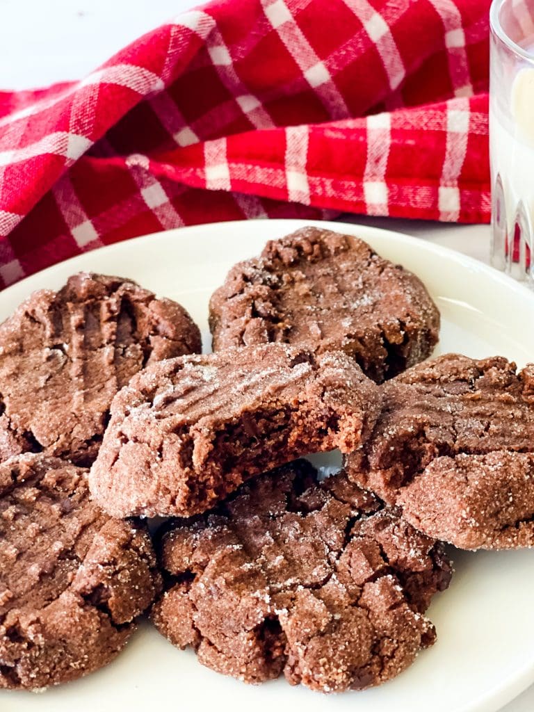 A plate of vegan chocolate peanut butter cookies on the white plate. The cookie on top has a bite taken from it. There is a red checked napkin behind and a glass of oat milk to the right.