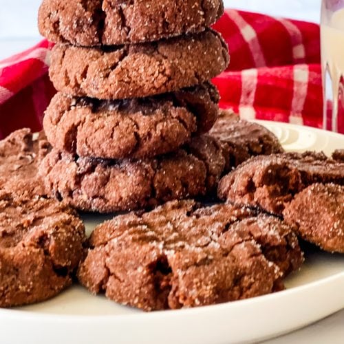 A stack of vegan chocolate peanut butter cookies with several on the white plate surrounding the stack. There is a red checked napkin behind and a glass of oat milk to the right.