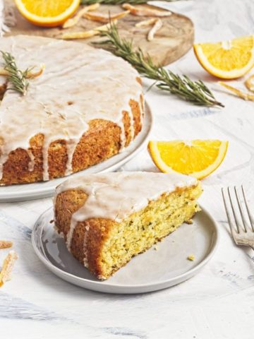 Vegan olive oil orange and rosemary cake on a white plate with orange halves and fresh sprigs of rosemary on top with a slice on a smaller plate and a fork.