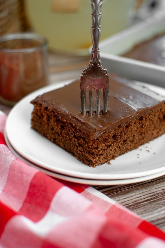 A square of vegan chocolate sheet cake placed on two white plates with a fork sticking in the top. There is a red checked napkin.