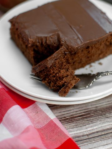 A square of vegan chocolate sheet cake placed on two white plates with a forkful removed. The fork is holding a piece of cake. There is a red checked napkin.