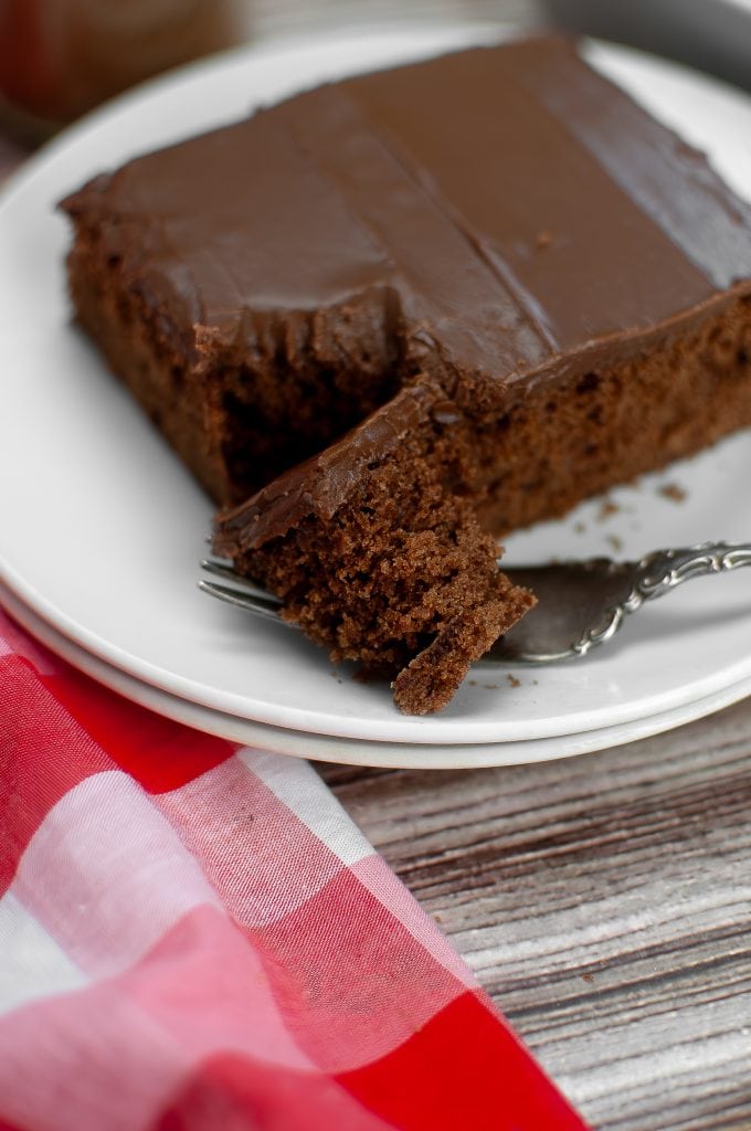 A square of vegan chocolate sheet cake placed on two white plates with a forkful removed. The fork is holding a piece of cake. There is a red checked napkin.