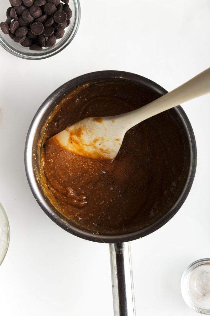 Stirring together the peanut butt4er and maple syrup to the small pan. A few bubbles on on the sides of the pan showing it is ready. Bowl of chocolate chips in top left corner.