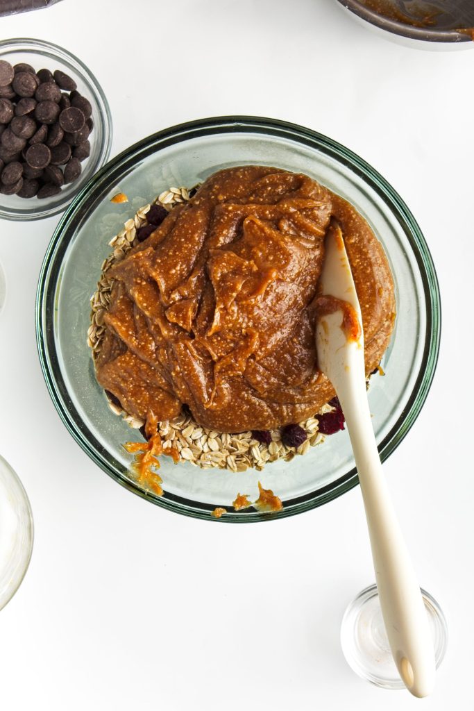 Adding the heated peanut butter mixture into the oat mixture. A bowl of chocolate chips in the top left corner.