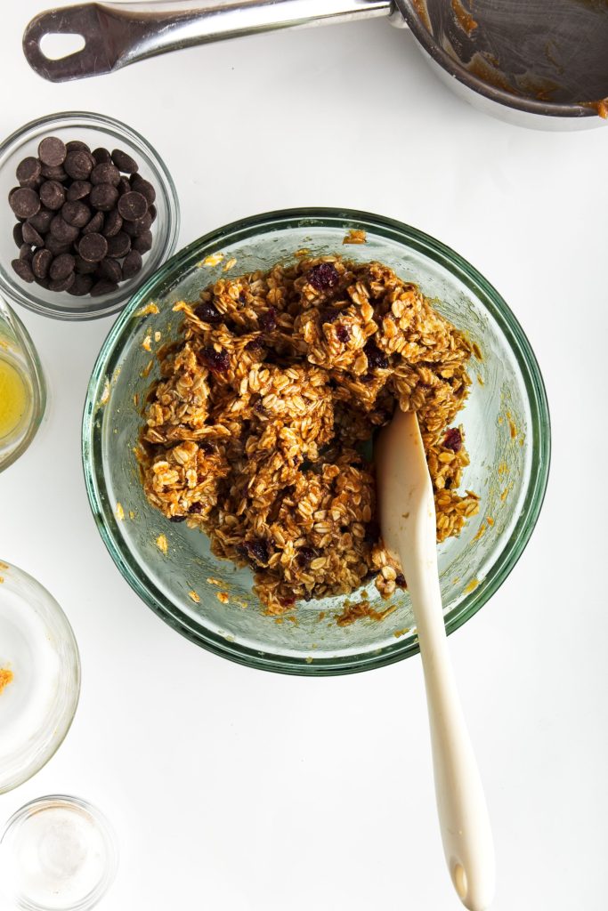 Stirring the heated peanut butter mixture into the oat mixture. A bowl of chocolate chips in the top left corner.