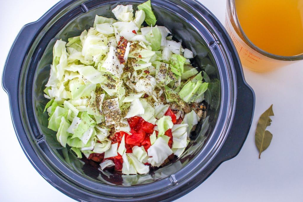 Vegetables in the crockpot with cabbage and seasonings on top. Veggie broth and bay leaf to the right side