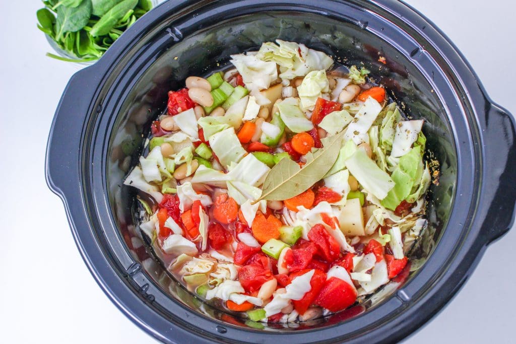 Vegetables in the crockpot with vegetable broth ready to cook. Bayleaf on top, cut spinach in the top left corner,