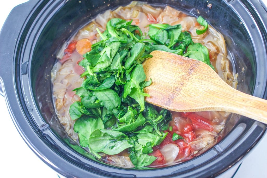 Stirring the spinach into the cooked minestrone soup in the slow cooker with a wooden spoon.