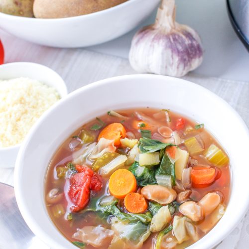 Minestrone soup in a white bowl. Spoon, vegan parmesan cheese, tomatoes, vegetables, garlic, and crockpot surrounding.