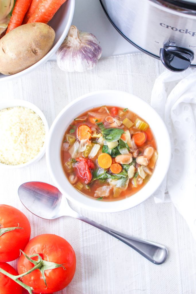 Minestrone soup in a white bowl. Spoon, vegan parmesan cheese, tomatoes, vegetables, garlic, and crockpot surrounding.