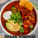 Bowl of vegan pumpkin chili with avocado, chips, jalapeno and sour cream on top with pinterest text overlay