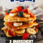stack of three ingredient vegan pancakes with bananas, blueberries, and cut strawberries on top,. All on a black plate with a gray plaid napkin in the background. with pinterest text overlay