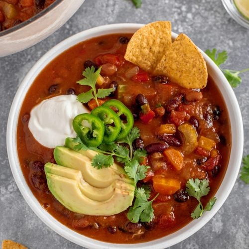 Hearty veggie pumpkin chili with avocado, vegan sour cream, and tortilla chips in a white bowl.