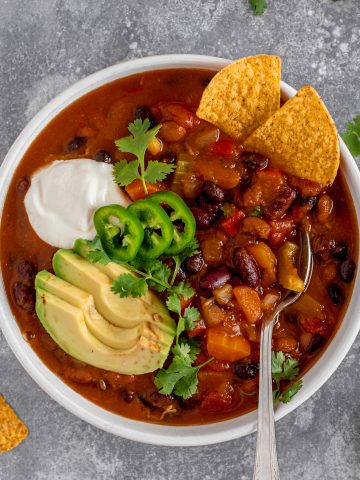 Hearty veggie pumpkin chili with avocado, vegan sour cream, and tortilla chips in a white bowl. Spoon in bowl,.