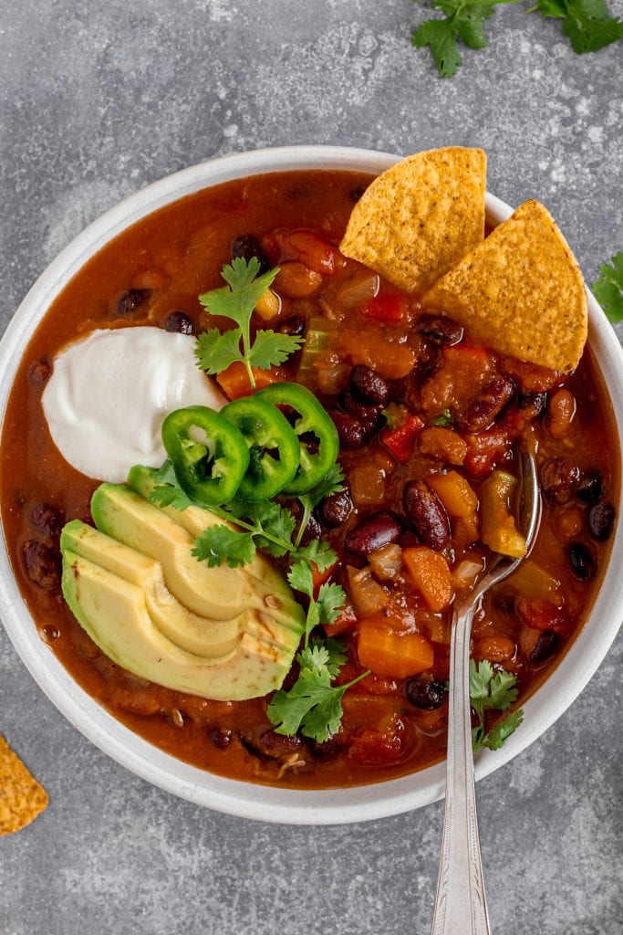 Hearty veggie pumpkin chili with avocado, vegan sour cream, and tortilla chips in a white bowl. Spoon in bowl,.