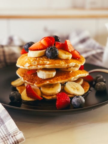stack of three ingredient vegan pancakes with bananas, blueberries, and cut strawberries on top,. All on a black plate with a gray plaid napkin in the background.