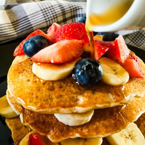 stack of three ingredient vegan pancakes with bananas, blueberries, and cut strawberries on top, with syrup being poured from a white pitcher. All on a black plate with a gray plaid napkin in the background.