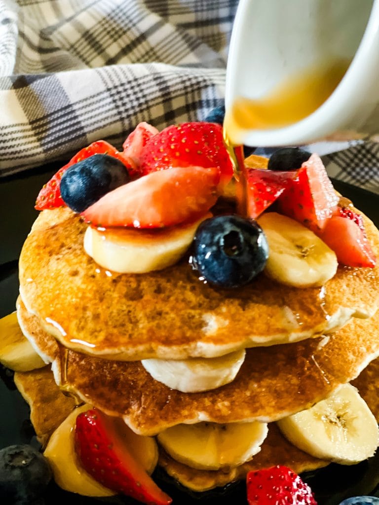 stack of three ingredient vegan pancakes with bananas, blueberries, and cut strawberries on top, with syrup being poured from a white pitcher. All on a black plate with a gray plaid napkin in the background.