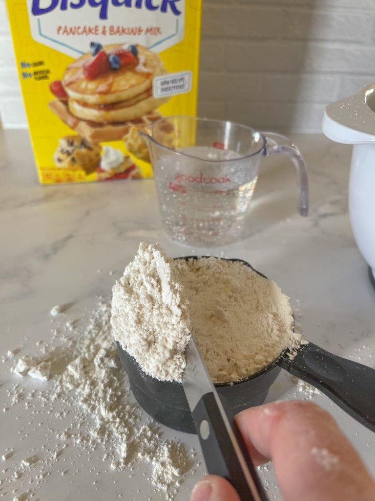Leveling off the Bisquick mix. A liquid measuring cup and a box of Biquick is in the background.