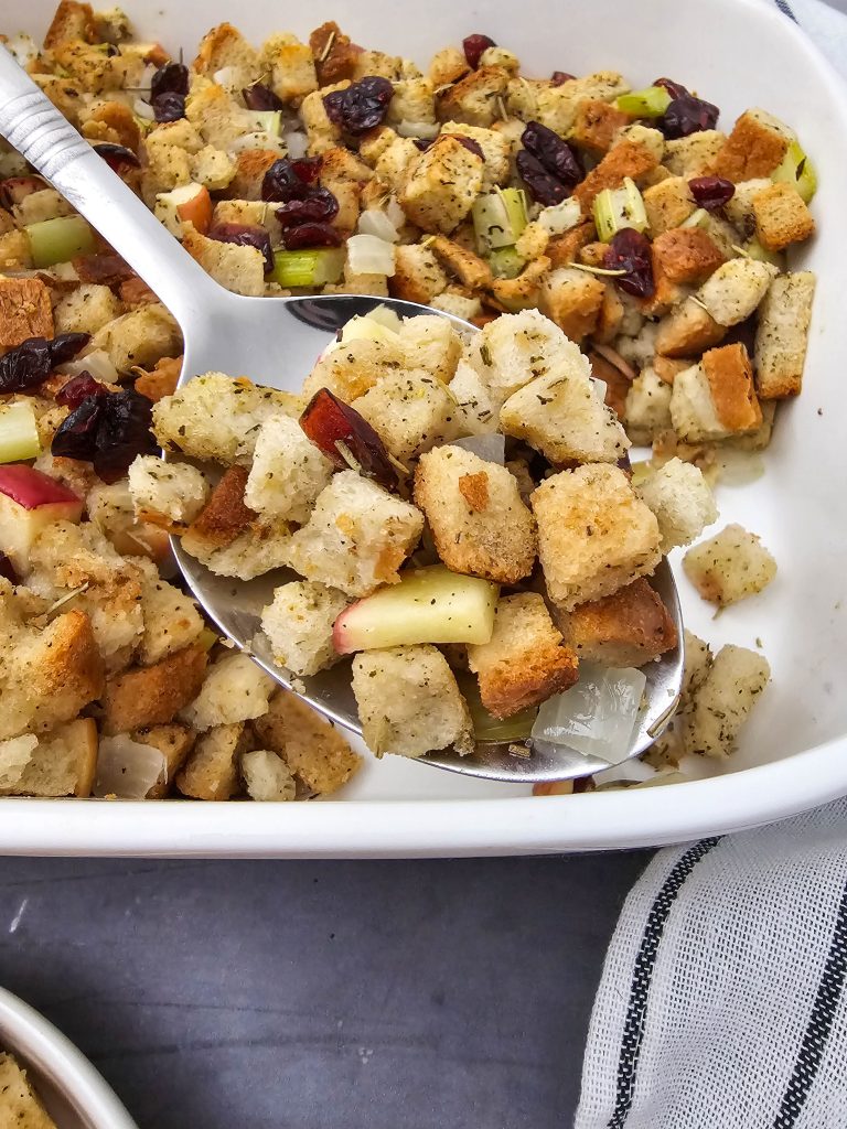 Spooning out vegan stuffing from a white casserole dish. Bits of apple and cranberry are showing.