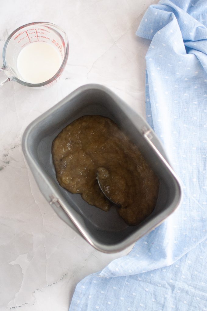 A bread machine pan with mashed bananas and a spoon inside, next to a glass measuring cup of plant milk and a light blue cloth on a marble countertop.