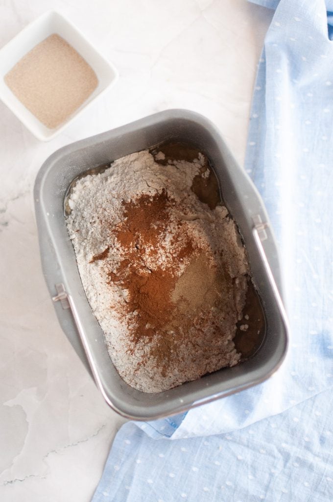 A bread machine pan filled with flour and various spices, ready for mixing. A small white dish with dry yeast sits nearby on a white surface, with a blue cloth partially visible to the side.