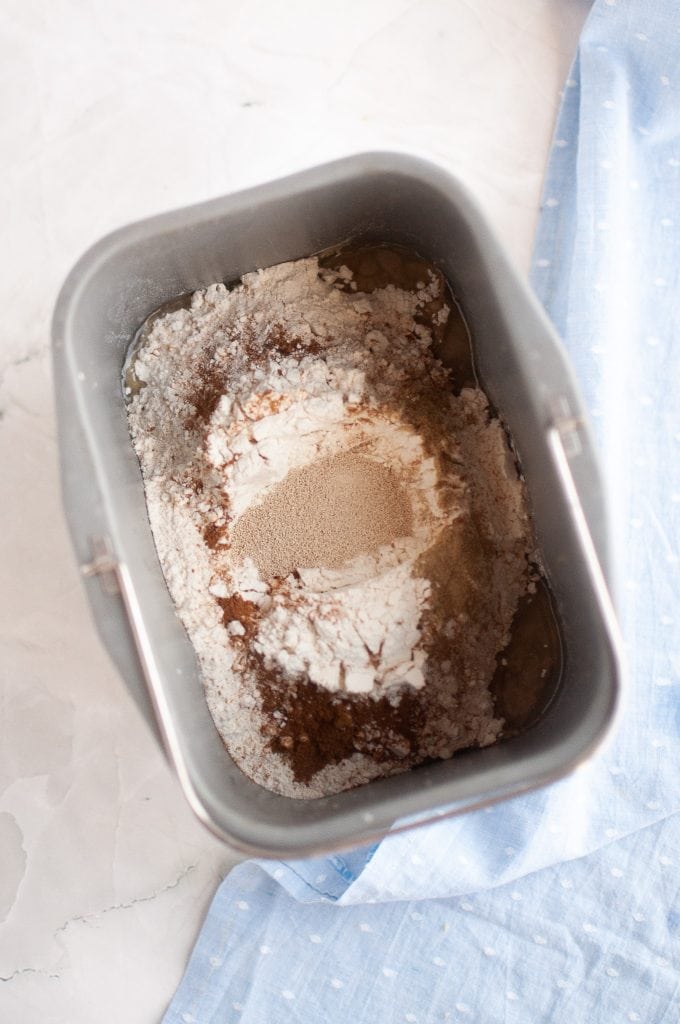 A bread machine pan filled with flour, yeast, and other dry ingredients, ready for baking, sits on a light-colored surface next to a blue cloth.