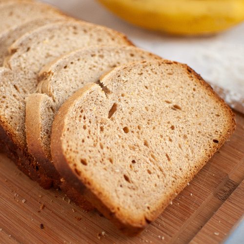 Four slices of homemade banana bread on a white plate, set on a light-colored surface next to a blue cloth and a partially visible banana.