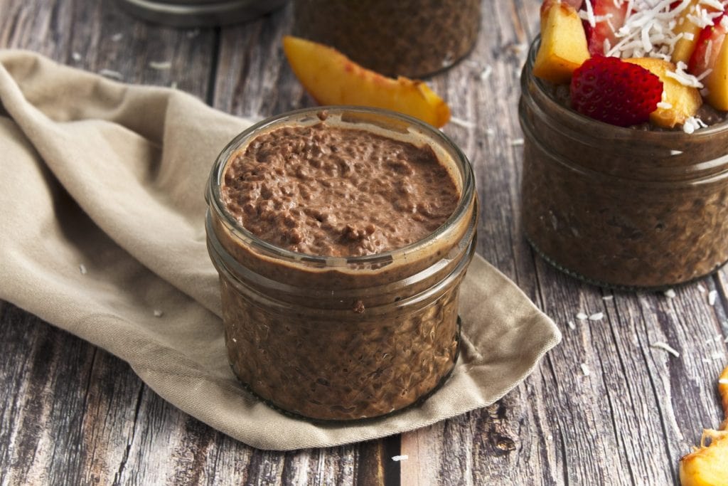 Chocolate chia Seed pudding in a jar topped with strawberries, peaches, an coconut shreds to the right and a plain non topped jar to the left