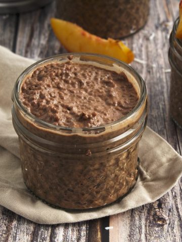 Chocolate chia Seed pudding in a jar topped with strawberries, peaches, an coconut shreds to the right and a plain non topped jar to the left
