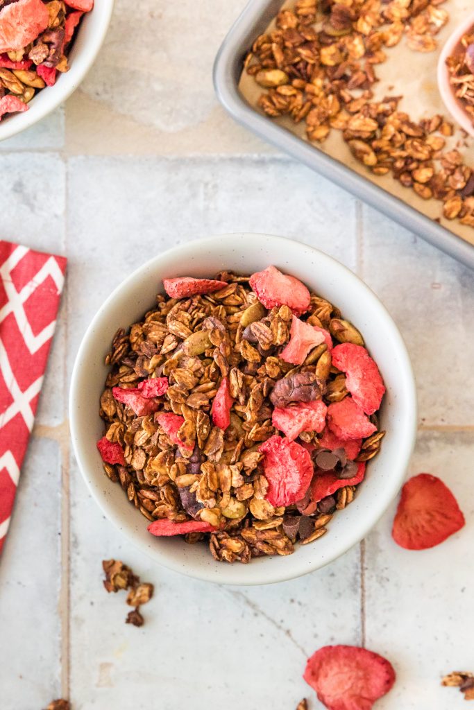 Strawberry chocolate granola in a white bowl with the sheet pan of granola in the background and a few strawberries and bits and pieces in the foreground