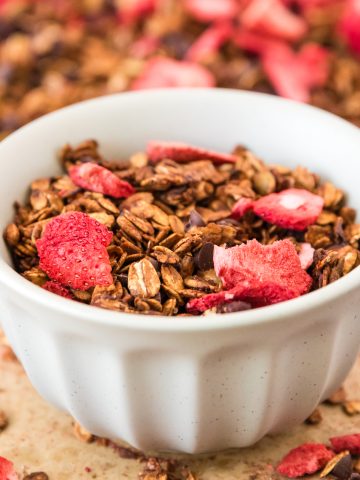 A white bowl of strawberry chocolate granola in the middle of a sheet pan full. The sections surrounding the white bowl has been cleared out.