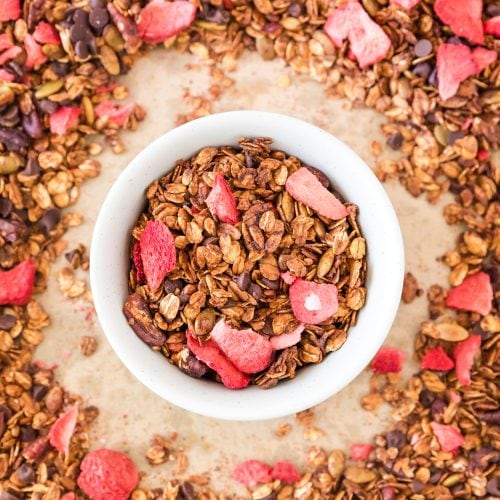 A white bowl of strawberry chocolate granola in the middle of a sheet pan full. The sections surrounding the white bowl has been cleared out.