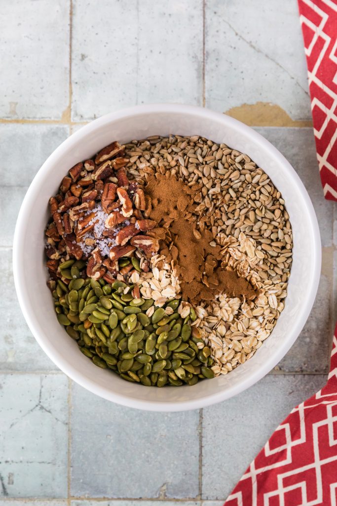 the dry ingredients for the strawberry granola in a white bowl waiting to be mixed together. One can clearly see the pepits, cinnamon and nuts on top of the oats,.