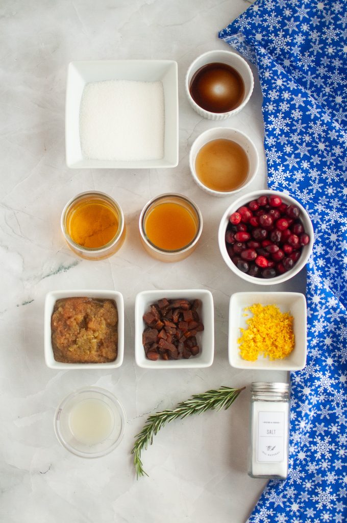 cranberry jam ingredients: Overhead view of various small bowls containing sugar, vanilla, liquids, cranberries, orange zest, fruit pieces, and spices on a marble surface, with a blue snowflake-patterned cloth in the corner.