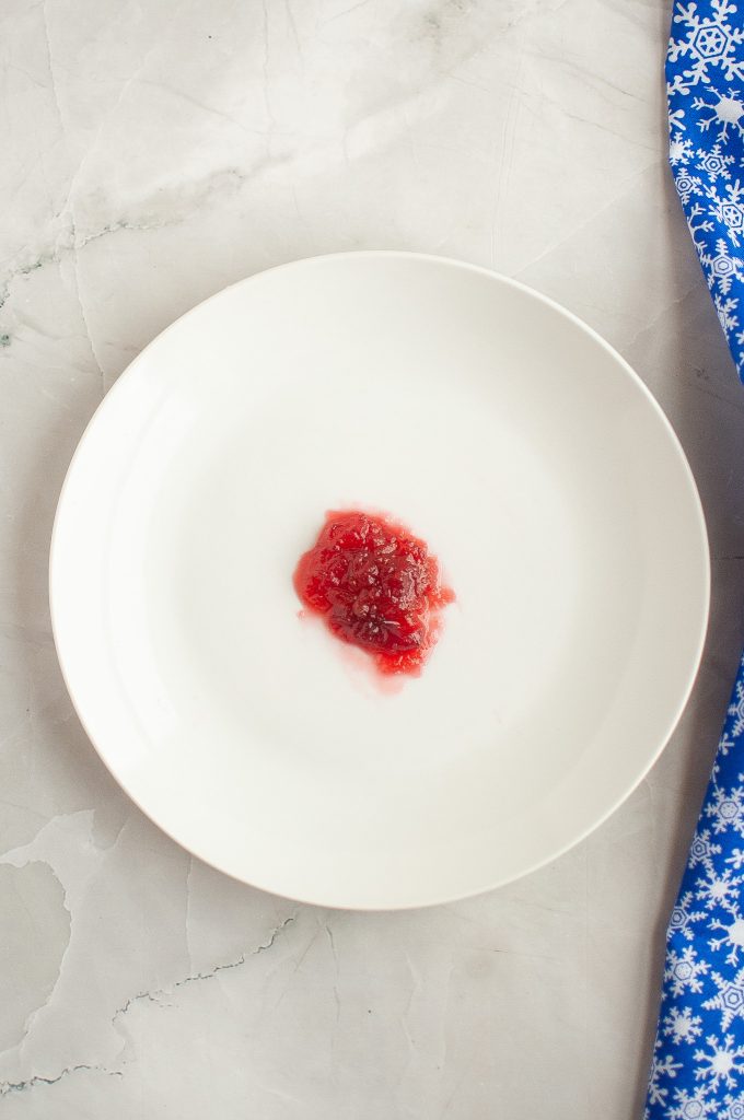 A small amount of red jelly or sauce is on a plain white plate, set on a light marble surface with a blue cloth featuring white snowflake patterns at the edge.