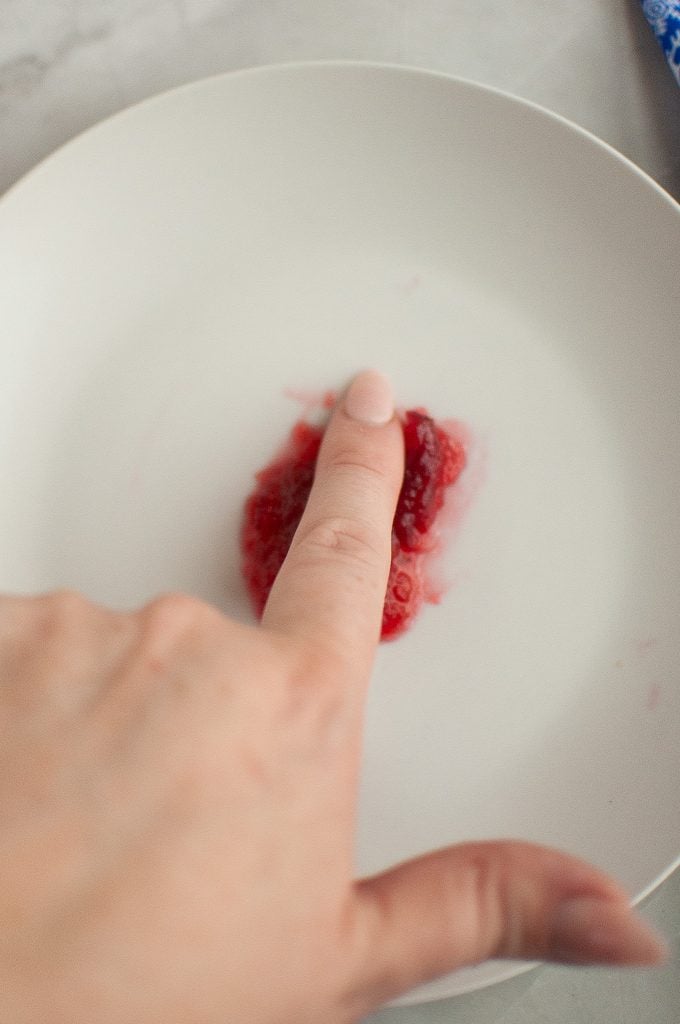 A hand presses a finger into a small pile of mashed raspberries on a white plate.