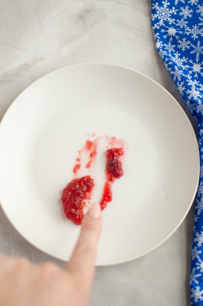 A finger points at a small smear of red jam or sauce on a white plate, with a blue and white snowflake-patterned cloth partially visible on the side.
