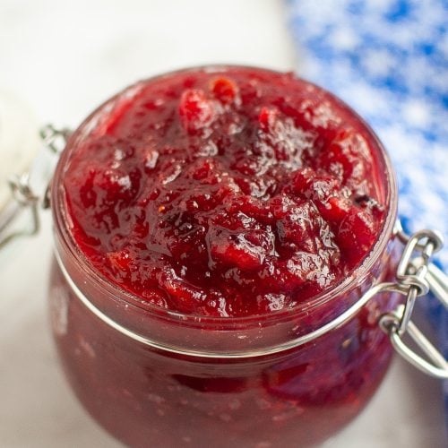 A glass jar filled with chunky, bright red cranberry sauce sits on a light surface with a blue and white patterned cloth blurred in the background.