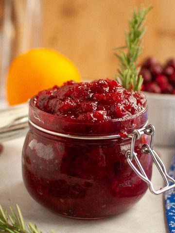 A glass jar filled with homemade cranberry jam sits on a white surface, garnished with a sprig of rosemary. In the background are cranberries in a white bowl, an orange, and a blue patterned cloth.
