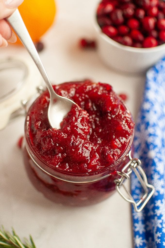 A spoon scooping homemade cranberry jam from a glass jar, with fresh cranberries and an orange in the background and a blue patterned cloth nearby.