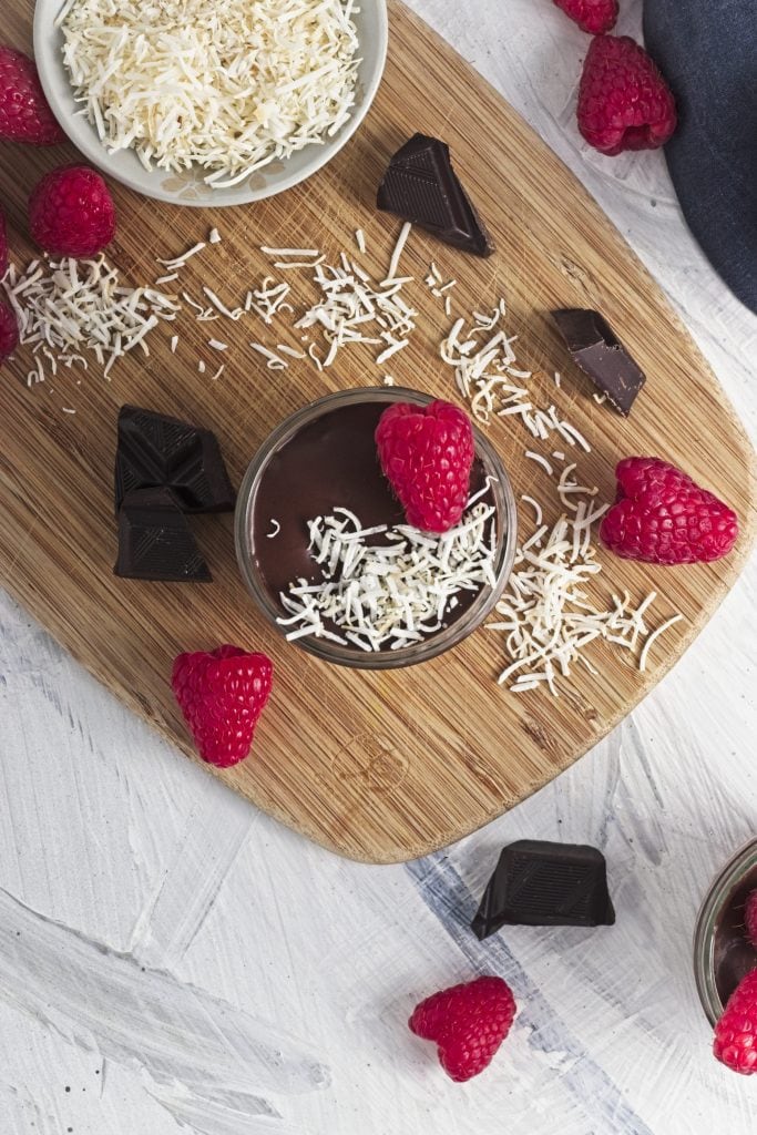 a wooden board with chocolate chunks, raspberries and coconut shreds with a bowl of shredded coconut and a jar of raspberry chia seed pudding with chocolate and coconut and a raspberry on top