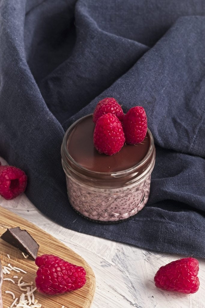 a wooden board with chocolate chunks, raspberries and coconut shreds with a bowl of shredded coconut and a jar of raspberry chia seed pudding with chocolate and coconut and a raspberry on top