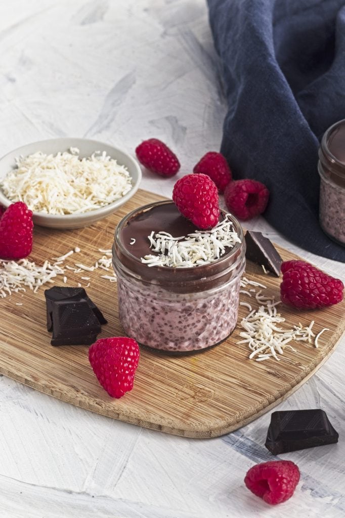 a wooden board with chocolate chunks, raspberries and coconut shreds with a bowl of shredded coconut and a jar of raspberry chia seed pudding with chocolate and coconut and a raspberry on top