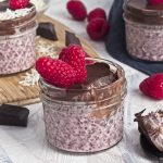 a wooden board with chocolate chunks, raspberries and coconut shreds with a bowl of shredded coconut and a jar of raspberry chia seed pudding with chocolate and coconut and a raspberry on top